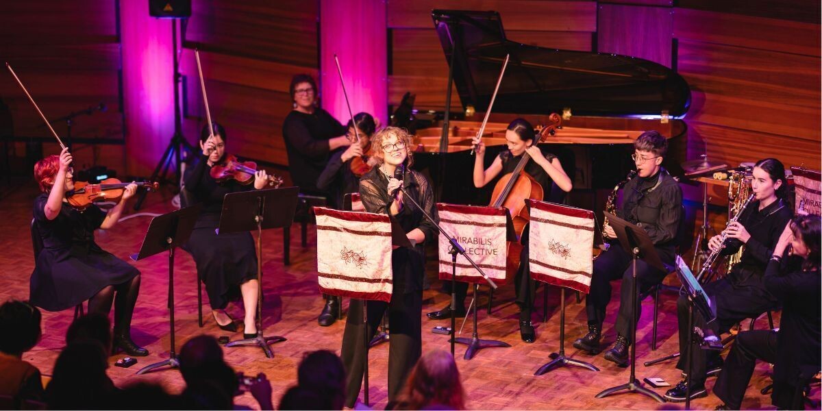 Members of Mirabilis Collective perform on stage under pink lighting. A singer stands at the microphone in front of musicians playing strings, woodwinds, and piano. Audience members watch as the ensemble performs with music stands draped in Mirabilis Collective banners.