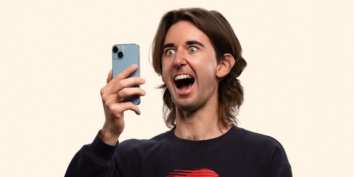 Fergus Neal - Boy–Comedian Fergus Neal with shoulder-length hair holds a smartphone in one hand, looking at it with wide eyes and an open mouth. He wears a dark sweatshirt with a red graphic on the front, set against a plain light-colored background.