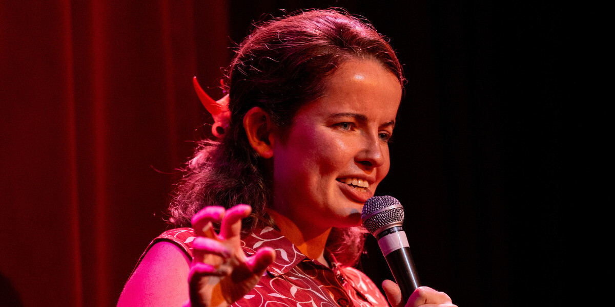 Roisin McAlinden, a woman with short brown hair, on stage speaking into a microphone. She looks confused and is making a strange gesture with her hand.