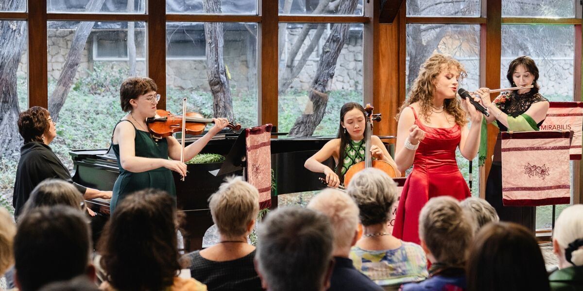 Five women musicians from Mirabilis Collective perform for an audience in a light-filled room. A pianist, violinist, cellist, and flautist accompany a singer in a red dress. Banners with the Mirabilis logo hang from the piano and music stand.