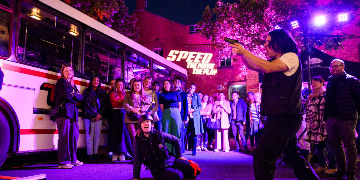 A crowd of people stand near an old-style bus. They watch several actors performing. One - dressed as a cop - is holding a prop gun towards the other, dressed as a villain. A third actor, also dressed as a cop, sits on the ground, having been shot.