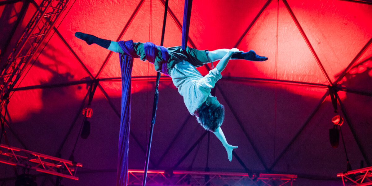 an aeiralist is hanging upside down with their legs in a split. they are shaded in blue while the background is shaded in red.