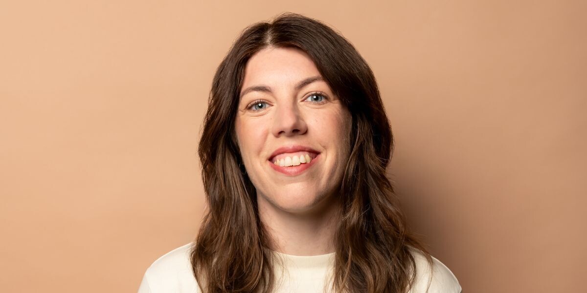 Alex Ward - Literally Anyone–Alex, smiling in front of peach toned background, she wears a white sweater