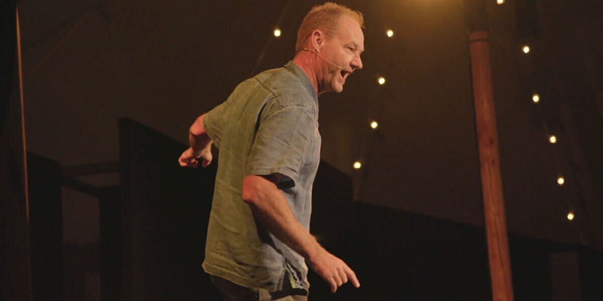 “Rob Carlton mid-performance on stage, speaking animatedly with stage lights glowing behind him.”