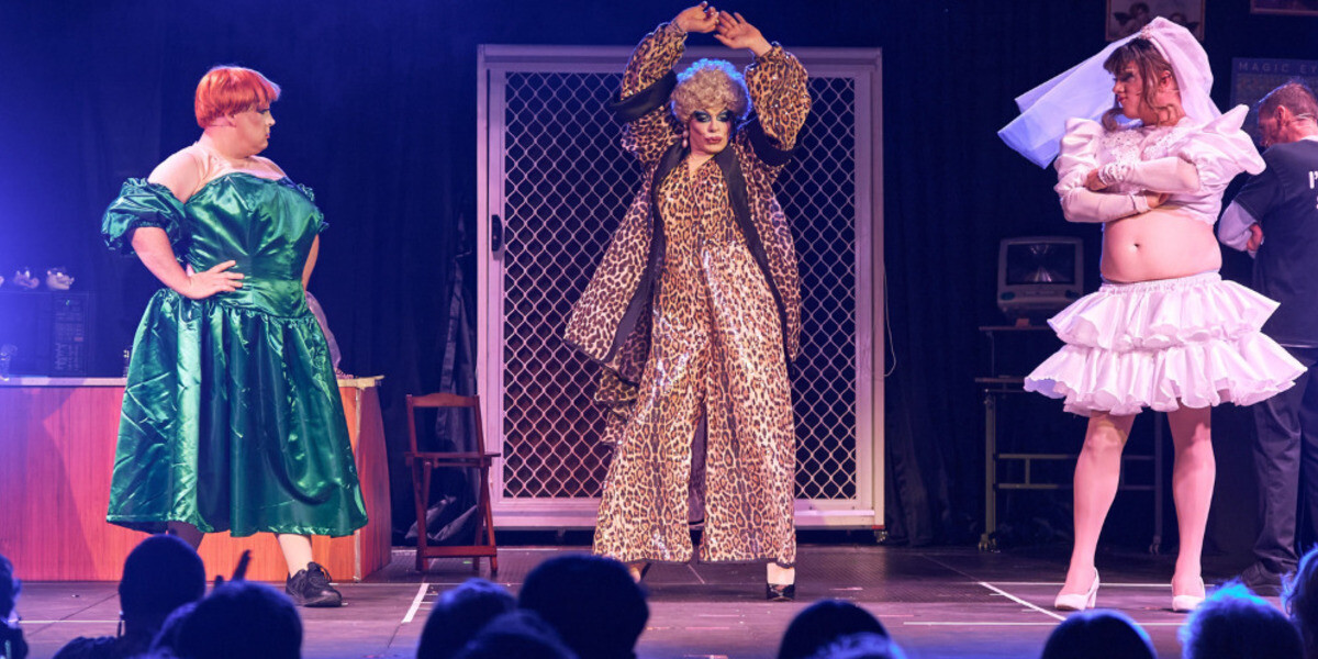 Two performers seated at a small café table on stage raise glasses in a toast. Left wears a bright pink two-piece with matching slides; right wears a red top with white trousers and a plush koala pinned to the chest. Blue mesh door backdrop and bottle on the table.