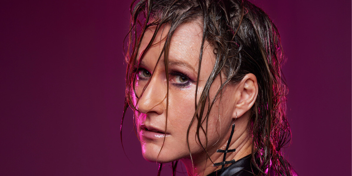 Image is of the performers face turned to the side, looking back at the camera. Her hair is oily and streaking all over her fac e She has a silver cross earring in.