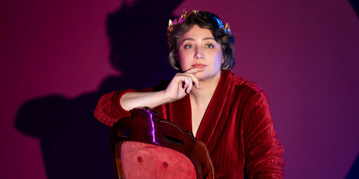 Laura Davis -Despair is Beneath Us–Laura Davis wearing a red velvet dressing gown and sitting on a red velvet chair a gold laurel crown on their head.