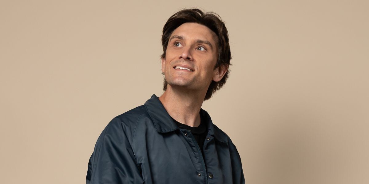 Sam Taunton - This Must Be The Place–Sam smiles while looking upwards, he is in front of a cream background, he wears a navy jacket.