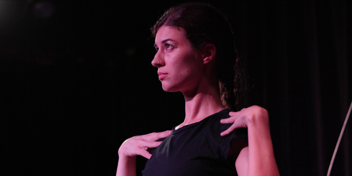 A profile shot of a woman on stage, hands resting on her shoulders, looking slightly frightened. She is wearing a black t-shirt.