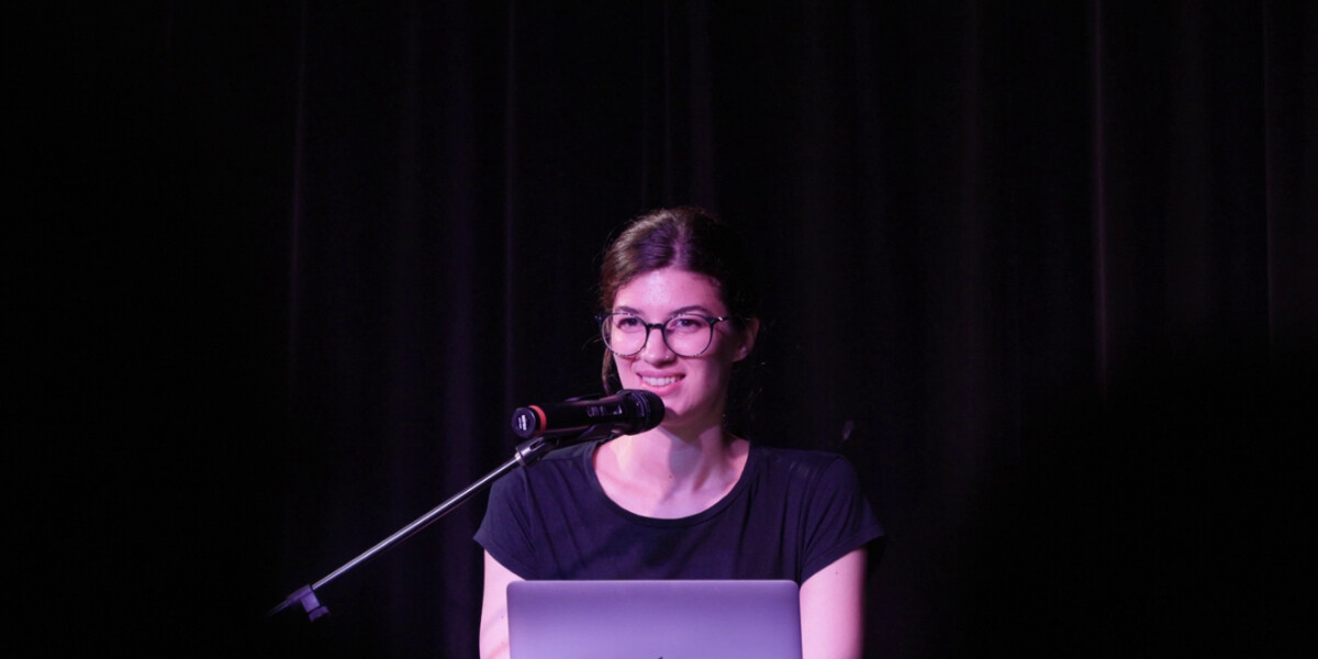 A woman on stage in front of a computer, speaking into a microphone on a stand. She is smiling. She is wearing glasses.