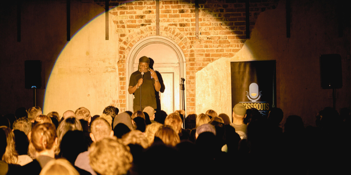 A packed audience watches a stand-up comedy performance at The Darkest Comedy Hour during FRINGE WORLD Perth. The comedian, Stephen K Amos, stands under a warm spotlight against a rustic brick wall, holding a microphone and engaging the crowd with laughter. A banner for Grassroots Comedy is visible beside the stage.