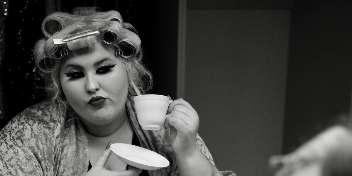 Charlee Watt sits at a makeup station in black-and-white styling, wearing hair rollers and a patterned robe. She holds a teacup and saucer in front of a mirror, with vintage accessories.