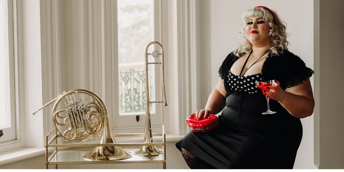 Charlee Watt: Beating Up The Beatles–Charlee Watt sits by a windowsill in a black polka-dot dress, holding a cocktail glass with a red cocktail and a red lip-shaped clutch. Beside her on a small stand are a trombone and French horn, with large windows and soft daylight behind her.