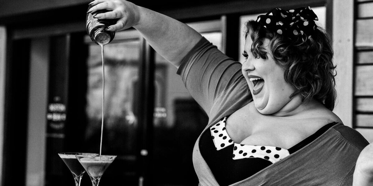 Charlee Watt stands outside Boston Bean Coffee Co., joyfully pouring a stream of espresso martini mix into two martini glasses on a bench.