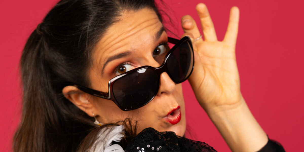 A close up of a woman in a sparkly black jacket with white angel wings and big black sunglasses. Looking down the camera and glasses at you. Pink background. Seductive and playful vibe.