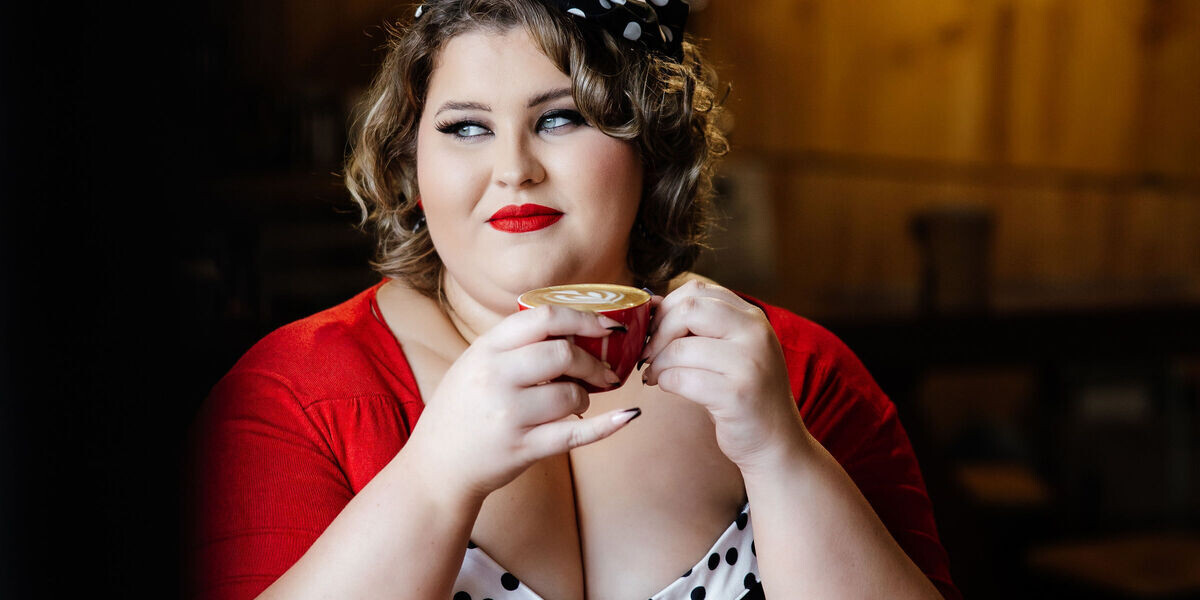 Charlee Watt: Caffeine Fiend - A Coffee Cabaret–Charlee Watt gazes to the side while holding a red coffee cup in a dimly lit wooden walled café. She wears a polka-dot headscarf and red bolero, evoking a vintage aesthetic.