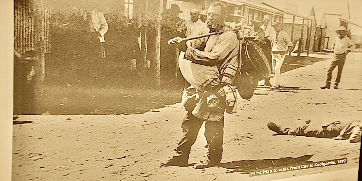 This is a Sepia image of a gold prospecting swagman standing in Bayley Street Coolgardie in the 1890s.
The swagman has a rolled blanket over his right shoulder and hanging to his front, while his left hand is holding a short pole laying over his left shoulder. Tied to the end of this pole are a number of what appear to be sundry supplies and small mining utensils.
The figure is bareheaded and is smoking a pipe.
In the background is an unpaved street lined with single story shopfronts and a small crowd men congregated along the shopfronts and edge of the street.
The caption reads "The first man to walk from Cue to Coolgardie 1892"