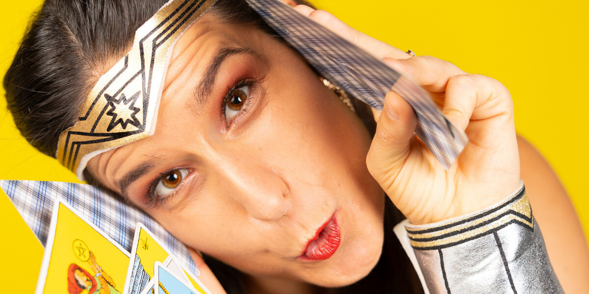 Psycho or Psychic–A close up of a woman in a Wonder Woman outfit, peering through tarot cards. A yellow background. An exciting, cheeky and playful vibe.