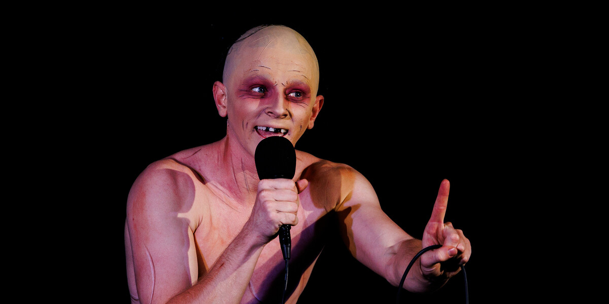 Image of character G'lem with a hand held microphone and wagging his finger at the audience.
