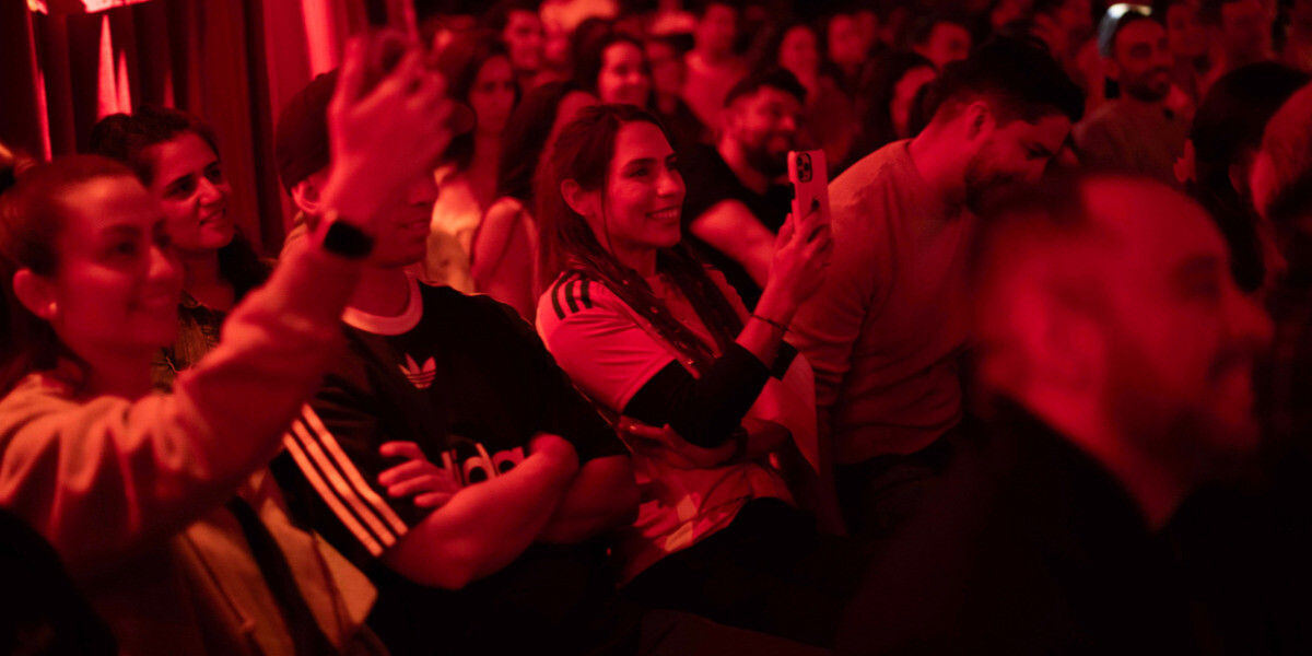 LATE ARVO LAUGHS–Audience members at a stand-up comedy show during FRINGE WORLD Perth, laughing and recording moments on their phones under warm red lighting. The crowd’s smiles and engagement reflect the energetic and interactive atmosphere of the night.