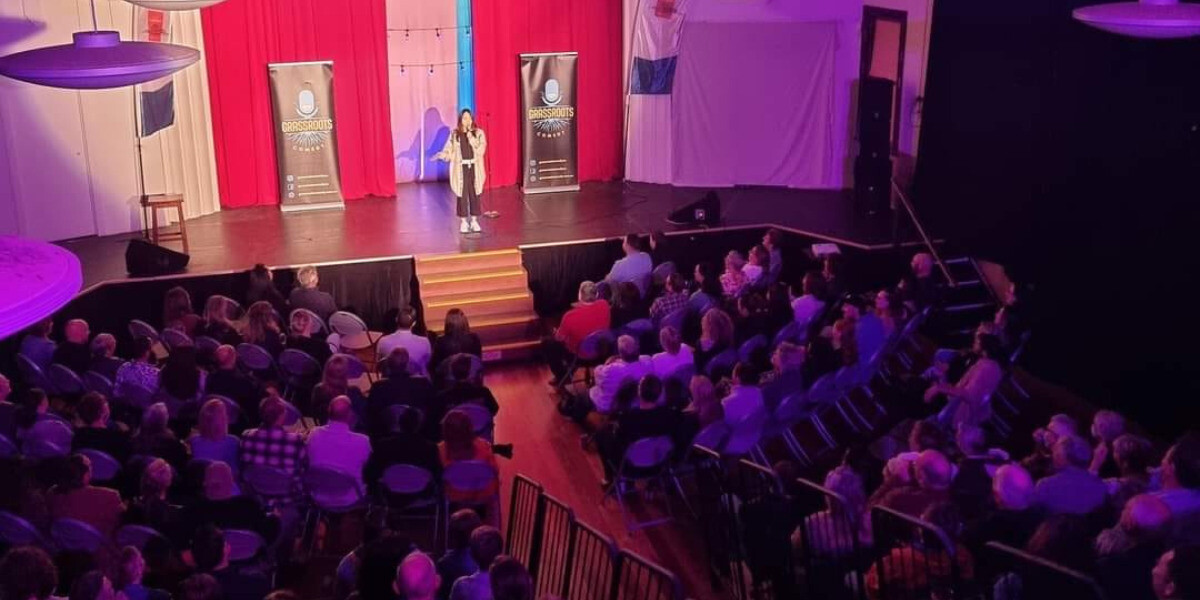 Wide-angle view of a stand-up comedy show presented by Grassroots Comedy during FRINGE WORLD Perth. A comedian performs on stage before a seated audience in a theatre with red curtains and warm lighting, creating an intimate, community atmosphere.