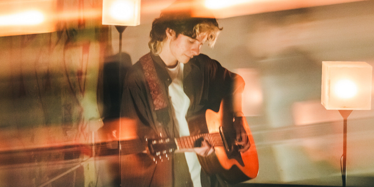 the worst of us.–a person with short brown hair with blonde sections is playing the guitar. the image has a blurred effect to it, and red-toned lights can be seen distorted around them. on the left is part of a graffiti wall.