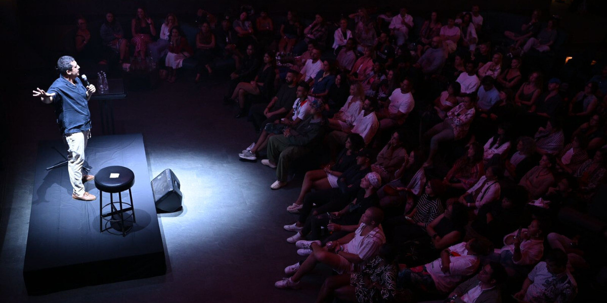 A stand-up comedian performs on a dimly lit stage in front of a packed audience. The performer stands beside a stool and a microphone stand, holding a microphone and gesturing mid-joke while the crowd watches attentively. A spotlight highlights the comedian against a dark background, creating an intimate club atmosphere.