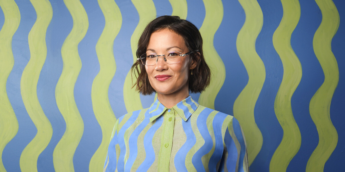 Lizzy Hoo - Says Hoo?–Lizzy smiles in front of a cream background, she wears a black shirt, glasses and red lipstick.