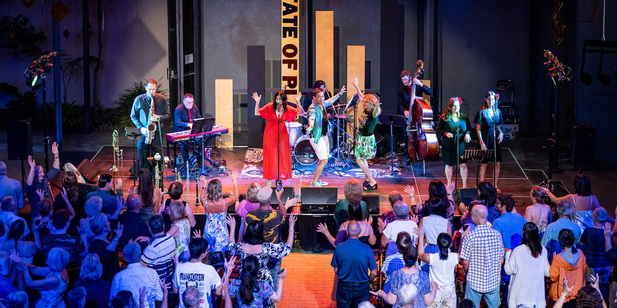 A jazz band featuring an Italian jazz singer in red and two backing vocalists perform on stage. Two dancers join them, while a big crowd dances, cheering and raising their hands.