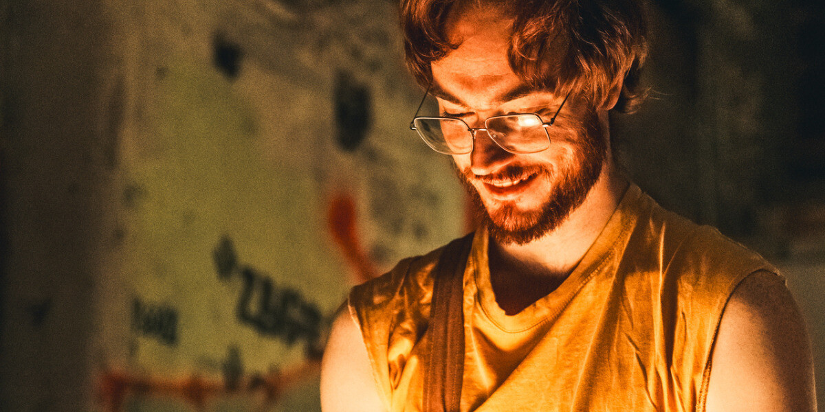 play.–a person with scruffy ginger hair and glasses looks towards the bottom of the image, where golden light emanates from an off-screen light source he is holding. he is wearing a yellow shirt and brown dungarees.