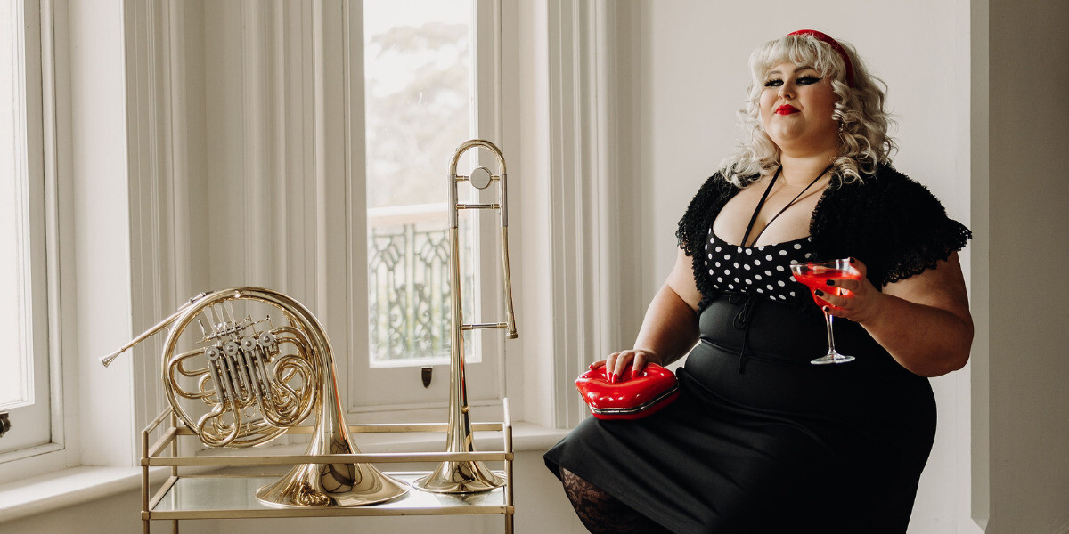 Charlee Watt sits by a windowsill in a black polka-dot dress, holding a cocktail glass with a red cocktail and a red lip-shaped clutch. Beside her on a small stand are a trombone and French horn, with large windows and soft daylight behind her.