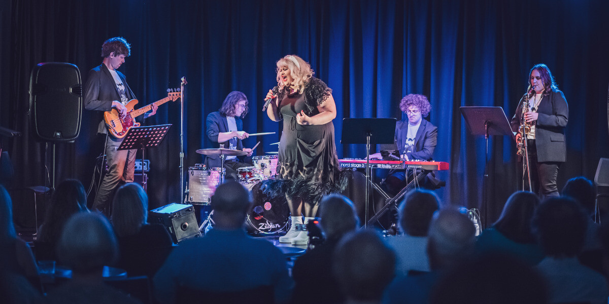 Charlee Watt performs on stage with her band, singing passionately into a microphone. She wears a black feather-trimmed dress while musicians on bass, drums, keys, and saxophone play alongside her. The audience is visible in the foreground, watching attentively.