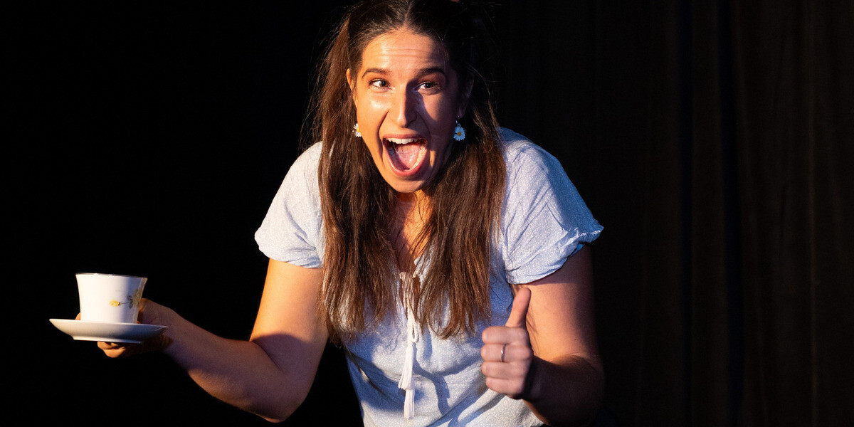 A woman holding a tea cup, with a big smile and a thumbs up looking at an audience member. Black background. Happy and affirming vibe.