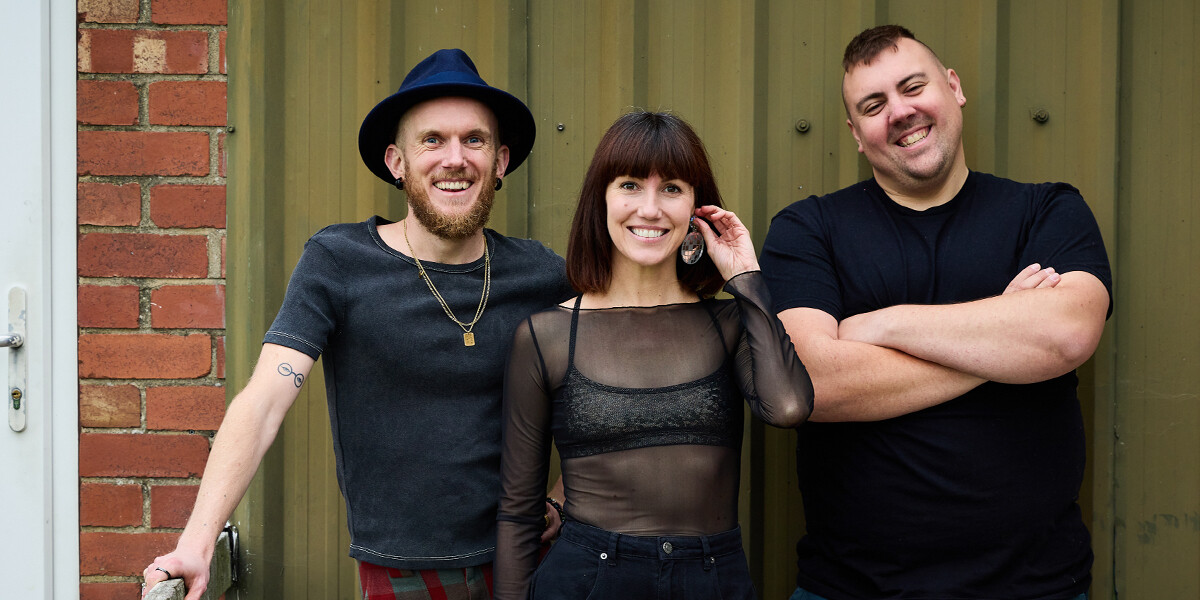 A writer in a hat, a performer in a sparkles, and a musician with folded arms, stood in front of a metal wall.