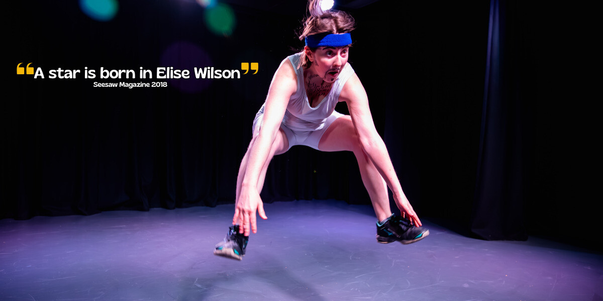 Performer in a pastel tracksuit and yellow headband stands on stage, holding out a black cap toward the audience. Stage lighting illuminates their face while audience members watch from the shadows.