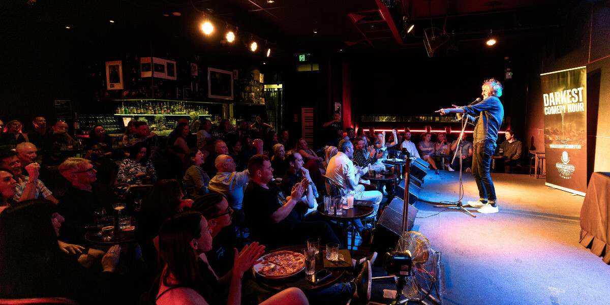 A lively crowd at The Darkest Comedy Hour during FRINGE WORLD Perth watches a comedian perform on stage at an intimate bar venue. The performer stands under bright stage lights with a microphone, while the audience laughs and applauds around tables with drinks and food. A banner for Grassroots Comedy appears beside the stage.