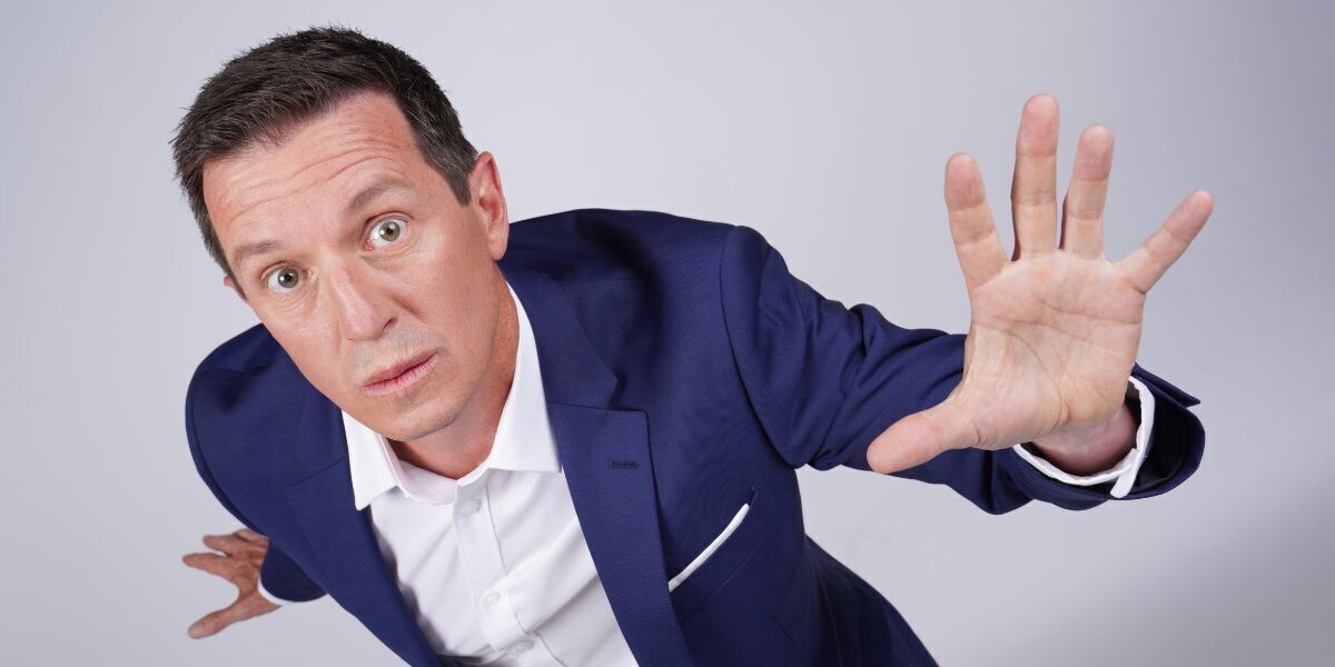 Tonight's Guest: Rove McManus (tales from the talk show trenches)–Comedian Rove McManus wears a blue suit jacket and white shirt, leaning forward with one hand extended toward the camera, fingers spread wide. He has short dark hair and looks directly at the camera with a surprised expression, set against a plain light gray background.