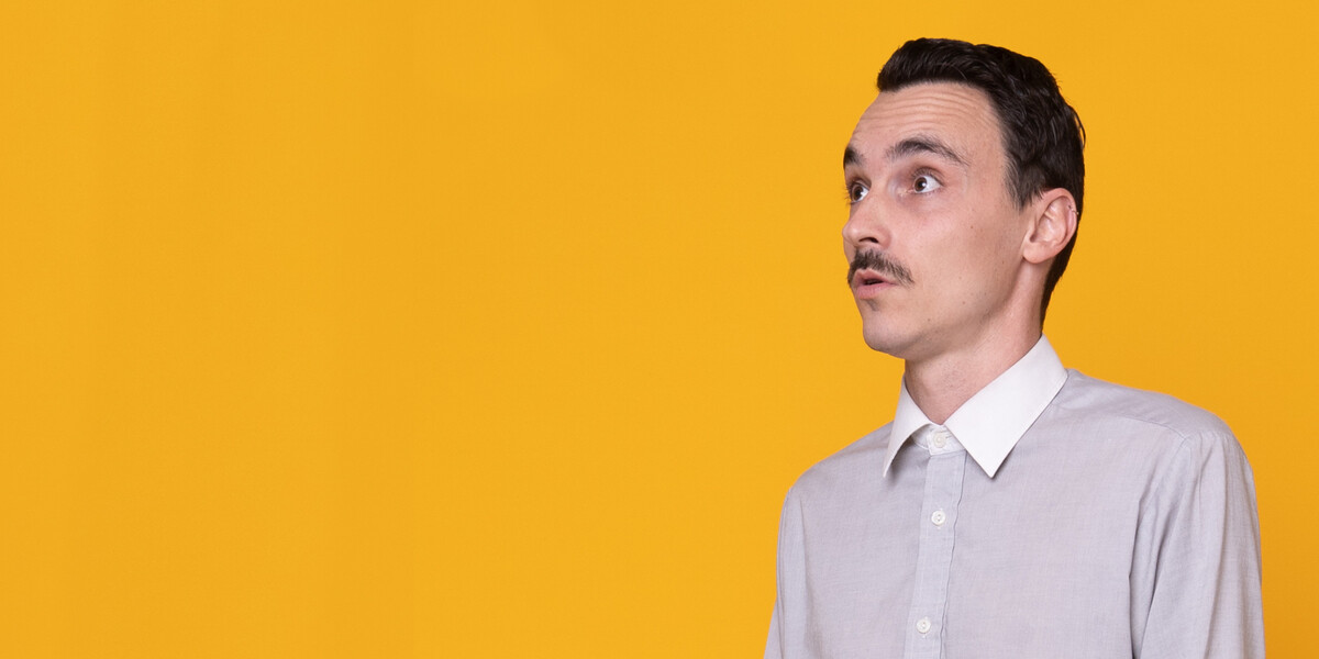 Full-body photo of a person standing against a bright yellow backdrop. They wear a light grey button-up shirt, dark trousers, and brown socks, standing with arms relaxed at their sides and looking slightly upward with a thoughtful expression.
