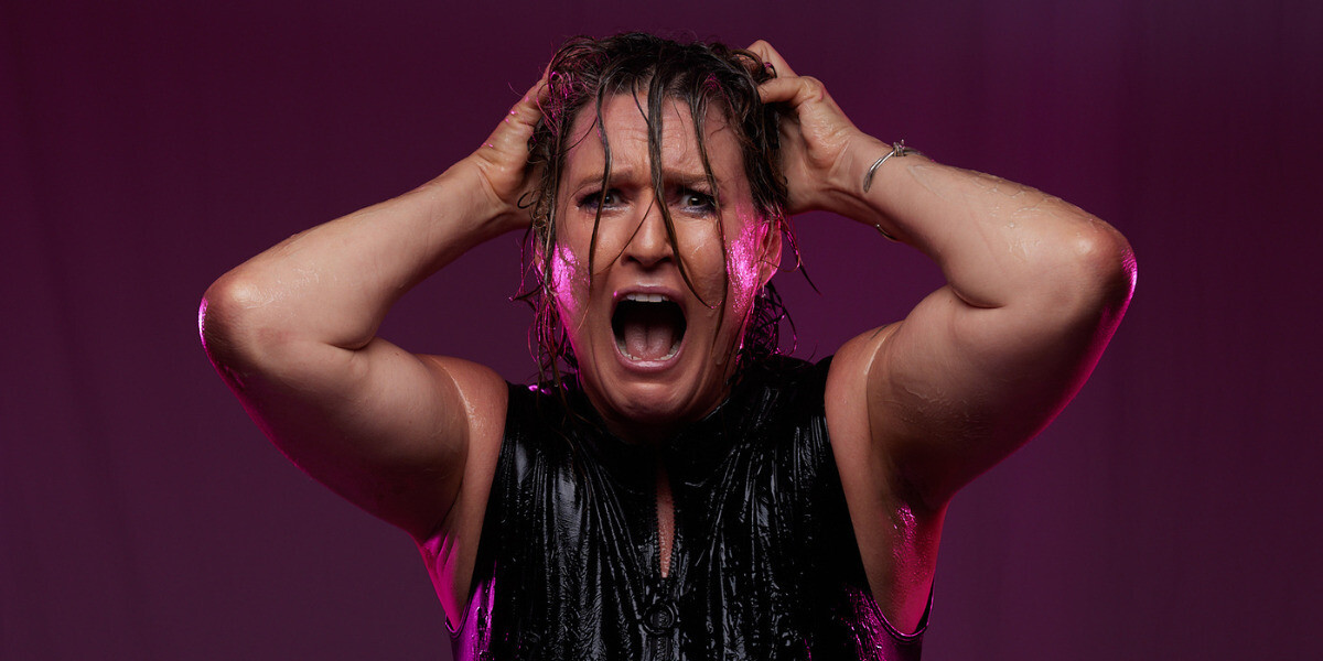 Image is of the performer with a scared, but comical look on her face. She is grasping at the hair on the top of her head with both hands. Her hair is all oily. Her outfit is a black, shiny body suit which is also oily.