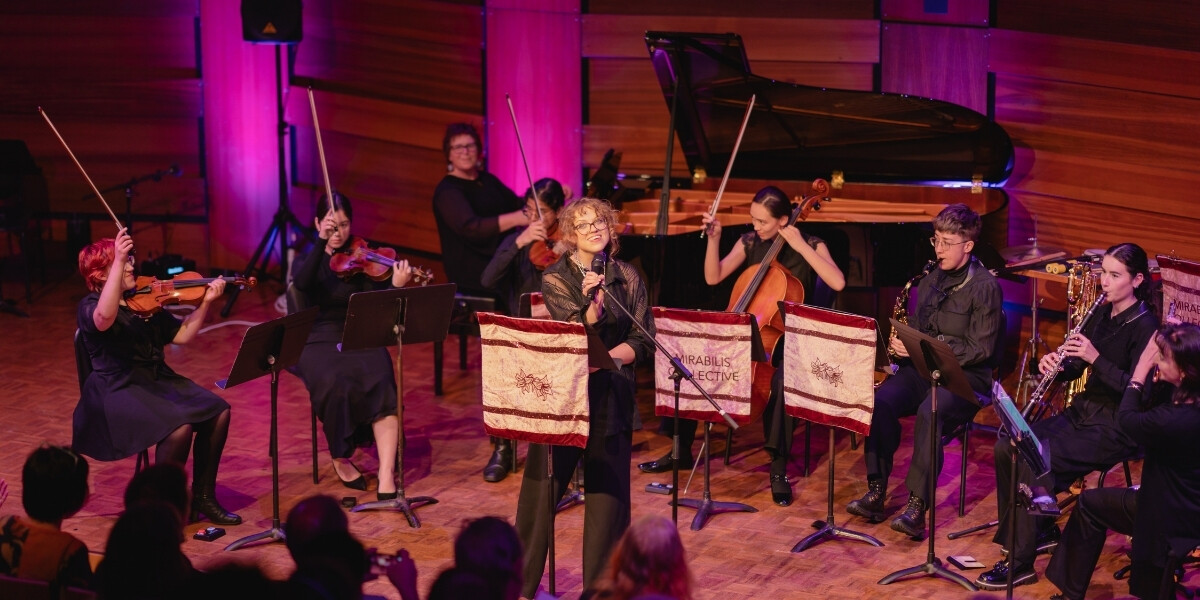 Mirabilis Collective performing on stage with strings, woodwinds, and piano. A vocalist stands at the centre microphone, smiling as the ensemble plays under warm purple lighting, with an audience watching in the foreground.