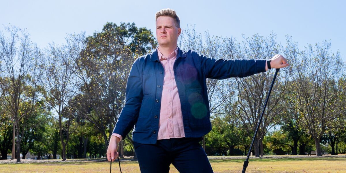Tom Ballard - Be Funny Challenge (Impossible)–Tom stand in a park, he holds a microphone and microphone stand, he is squinting from the sun.