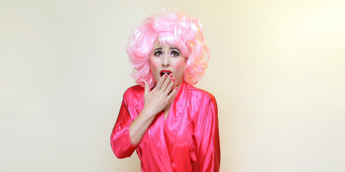 Lucy Best: Unbelievable–MUAH/Styling and Photography by Rachel Mia
Lucy Best is wearing a pink wig and dress, she has one hand to her mouth in a mock shock look. It's very camp.