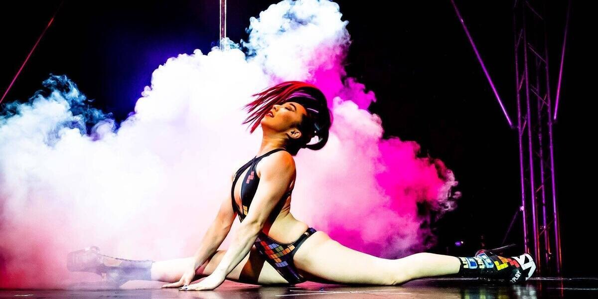 Asian woman wearing Tetris themed bodysuit & platform stiletto boots does a split while flipping her long colourful ponytail and surrounded by smoke