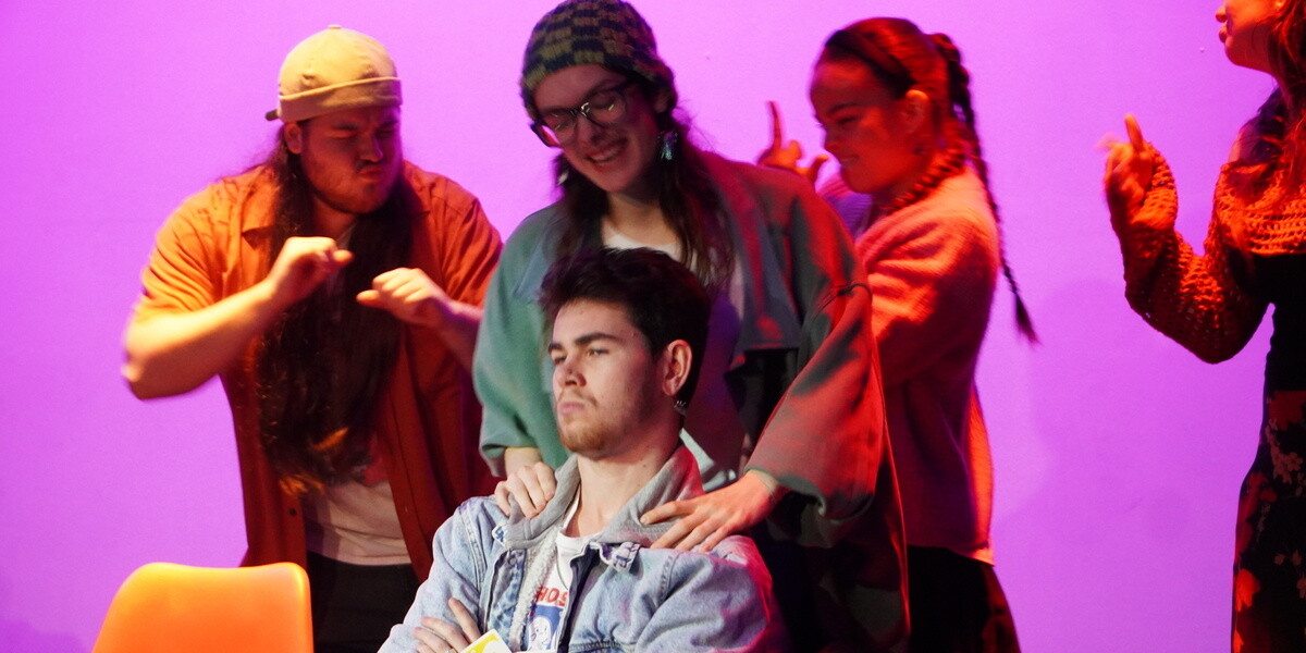 Four actors dancing around a male actor, sitting in a chair.