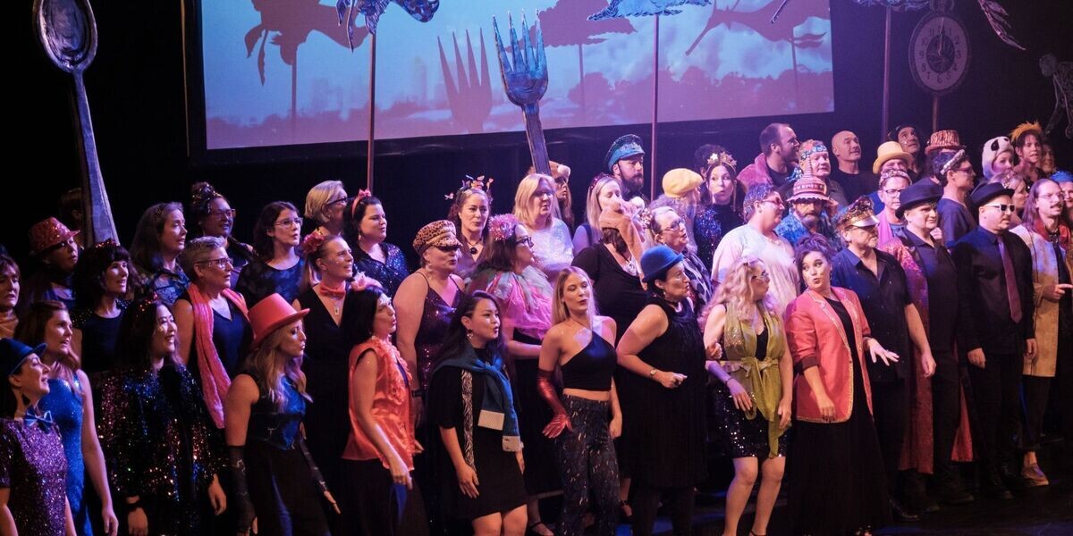 A big bank of grooving choristers in colourful and arty outfits sing their hearts out. Behind them are larger than life props on sticks - fish, spoon, fork, clock.