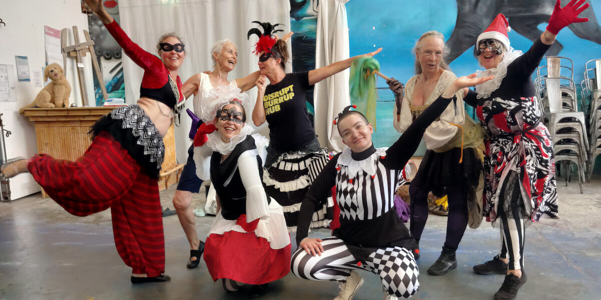 Fossil Fool Crime Walk - Street theatre tableau outside a stone building: a crowned “king” in red robes sits on a chair, a masked figure stands beside a wooden gallows frame holding ropes, and a robed “judge” sits at a red-draped table beneath a banner.