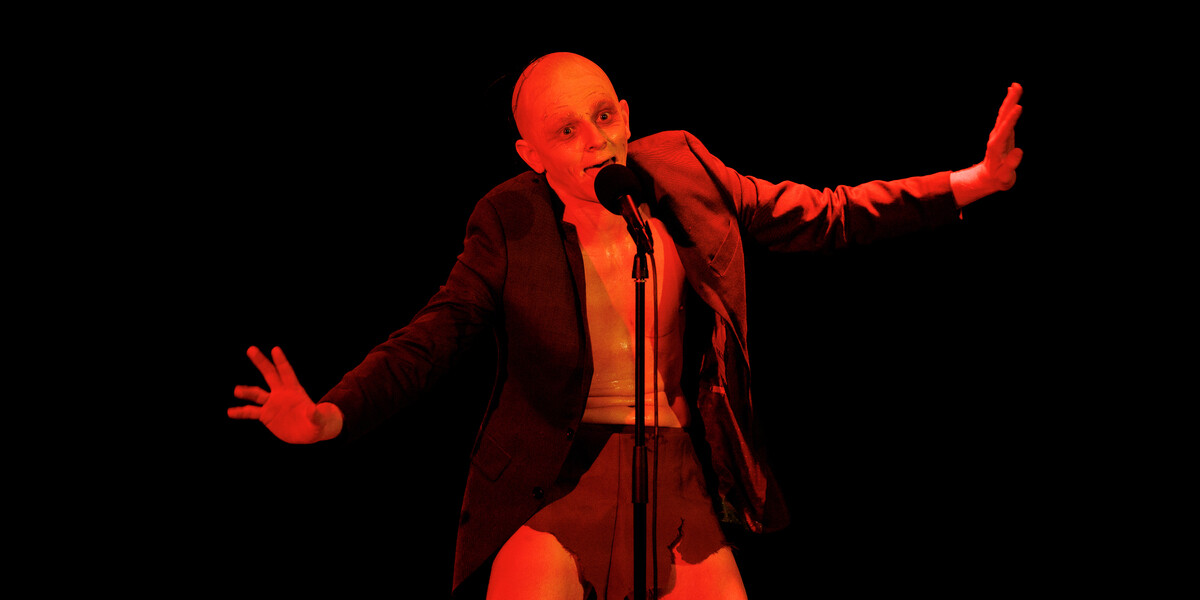 Image of character G'lem on stage awash in a nightmarish red light, singing into the microphone wearing a grey suit jacket.