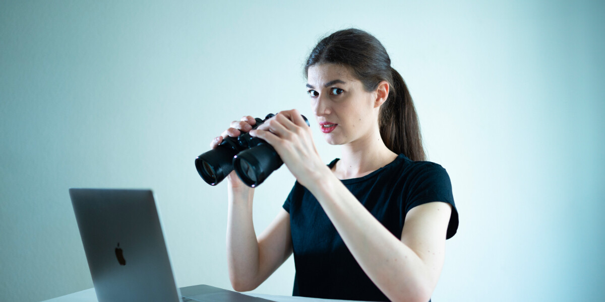 Embarrassed Naked Female (this show contains nudity)–A woman sitting at a desk facing a computer, holding binoculars in her hands. She looks at the camera with a slightly uneasy expression. She wears a black t-shirt and has her hair in a ponytail.