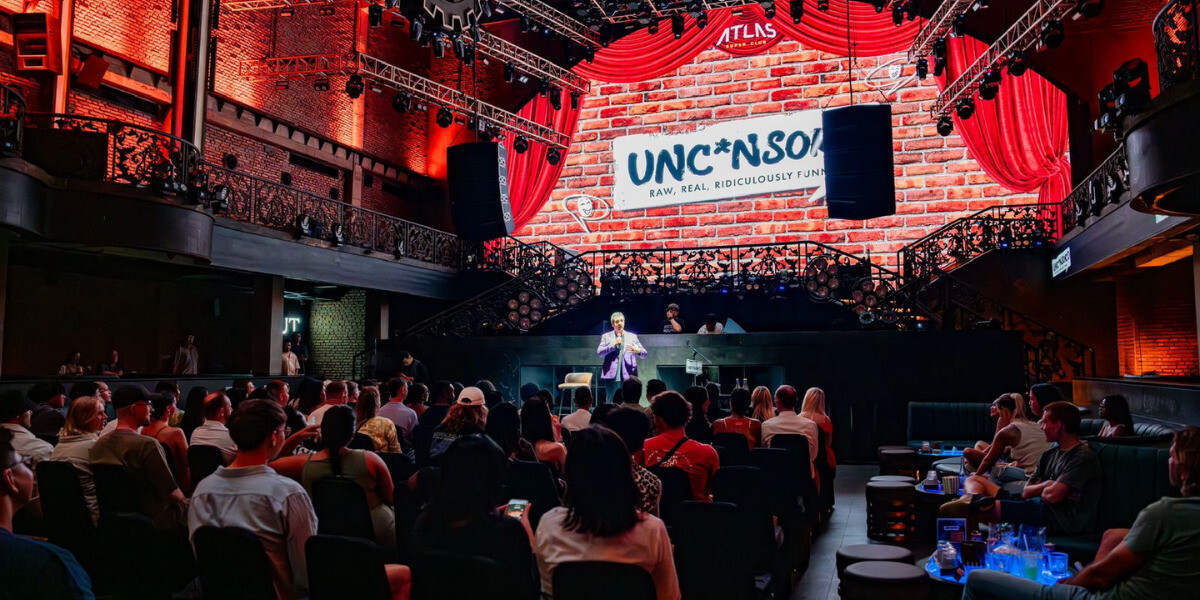 A stand-up comedy show takes place inside a grand, modern theatre with red curtains and industrial-style balconies. The stage features a large screen displaying the word “UNC*NSORED” with the tagline “Raw, Real, Ridiculously Funny.” The comedian stands under bright lights, addressing a lively audience seated at tables and lounge chairs throughout the venue