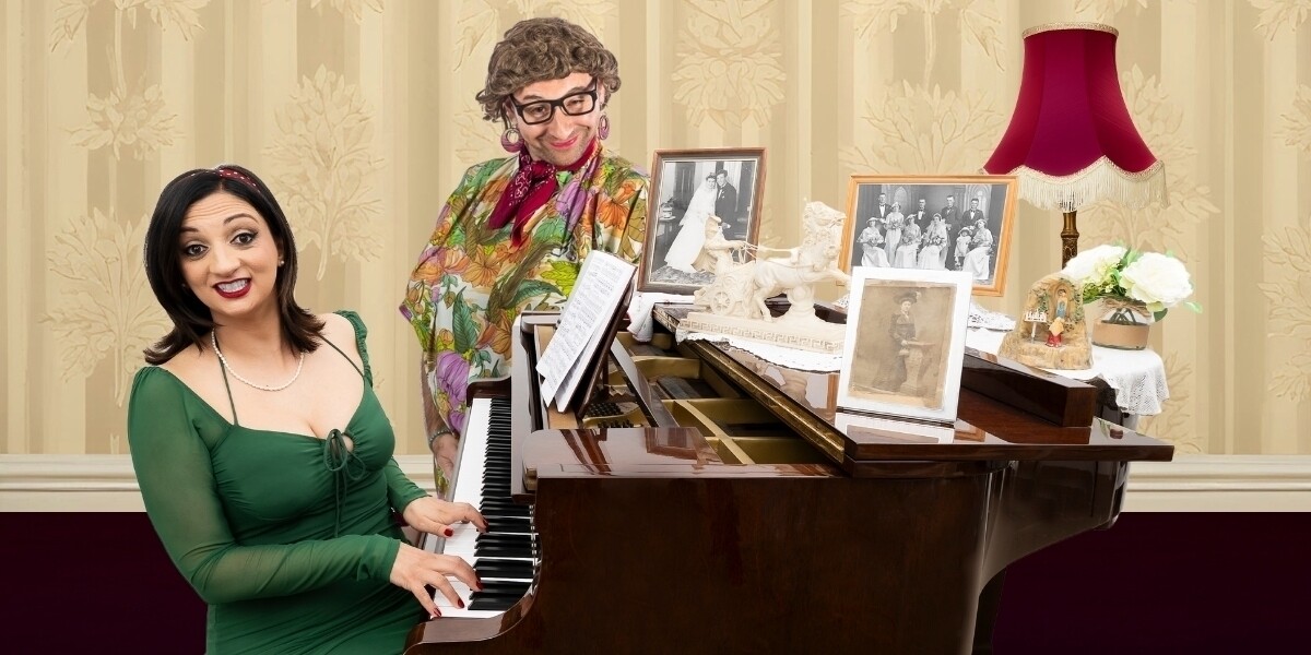 NonnaMia!–An Italian woman sits at a piano with her smiling nonna behind her, surrounded by black-and-white photos, cream wallpaper, and a retro red lamp.